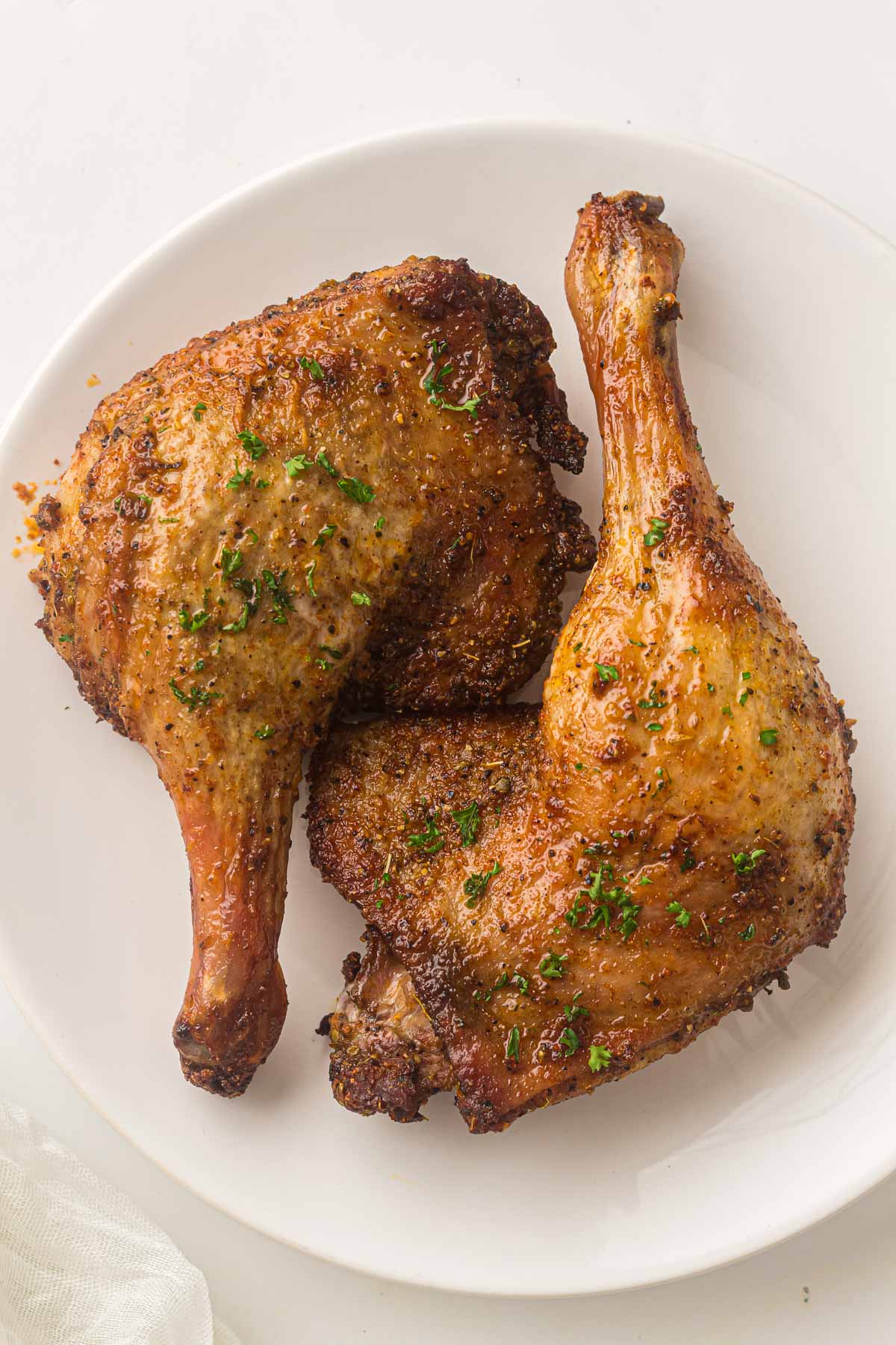 Two cooked and seasoned duck leg quarters with crispy skin served on a white plate, garnished with chopped herbs.