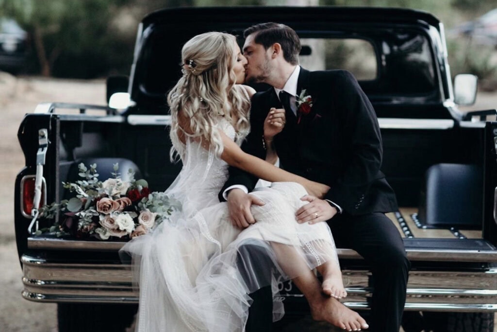 A bride and groom sit in the back of a pickup truck and share a kiss