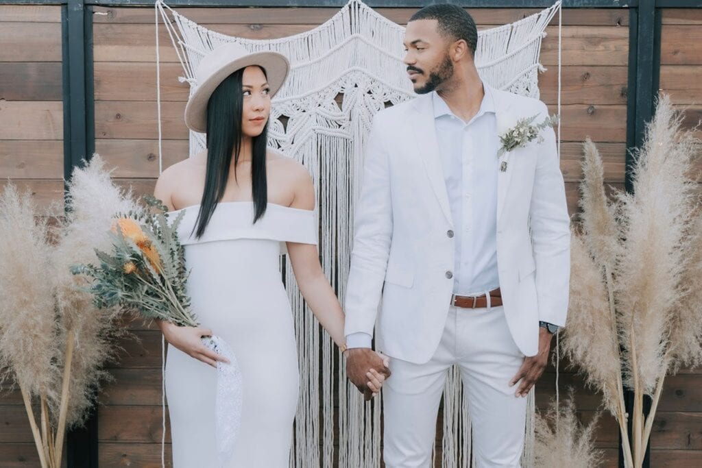 A bride and groom in all white stand hand in hand at a bohemian-inspired altar