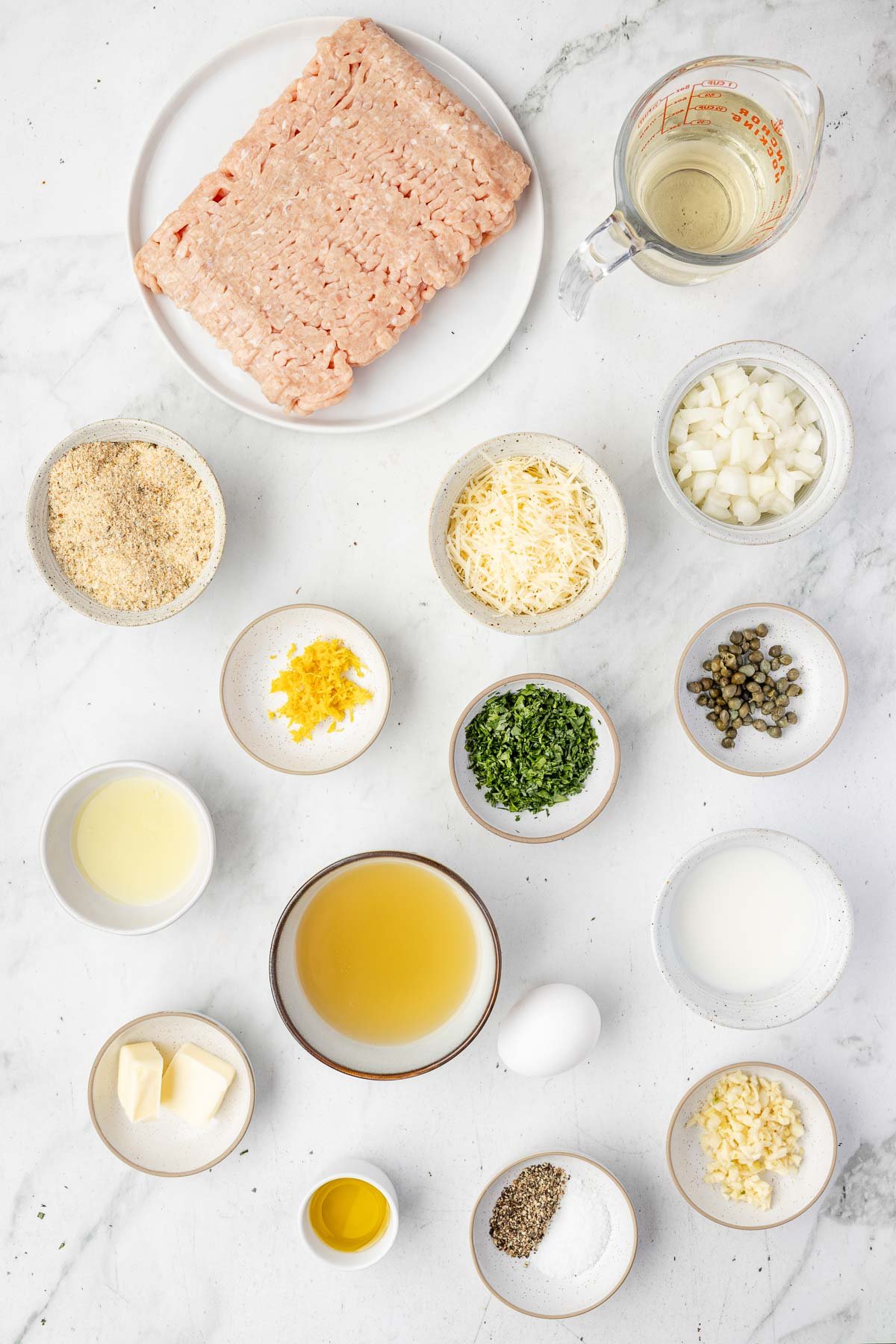 Ingredients for Chicken Piccata Meatballs displayed on a white surface, including ground meat, chopped onions, bread crumbs, shredded cheese, herbs, capers, lemon zest, an egg, broth, butter, oil, milk, and seasonings.