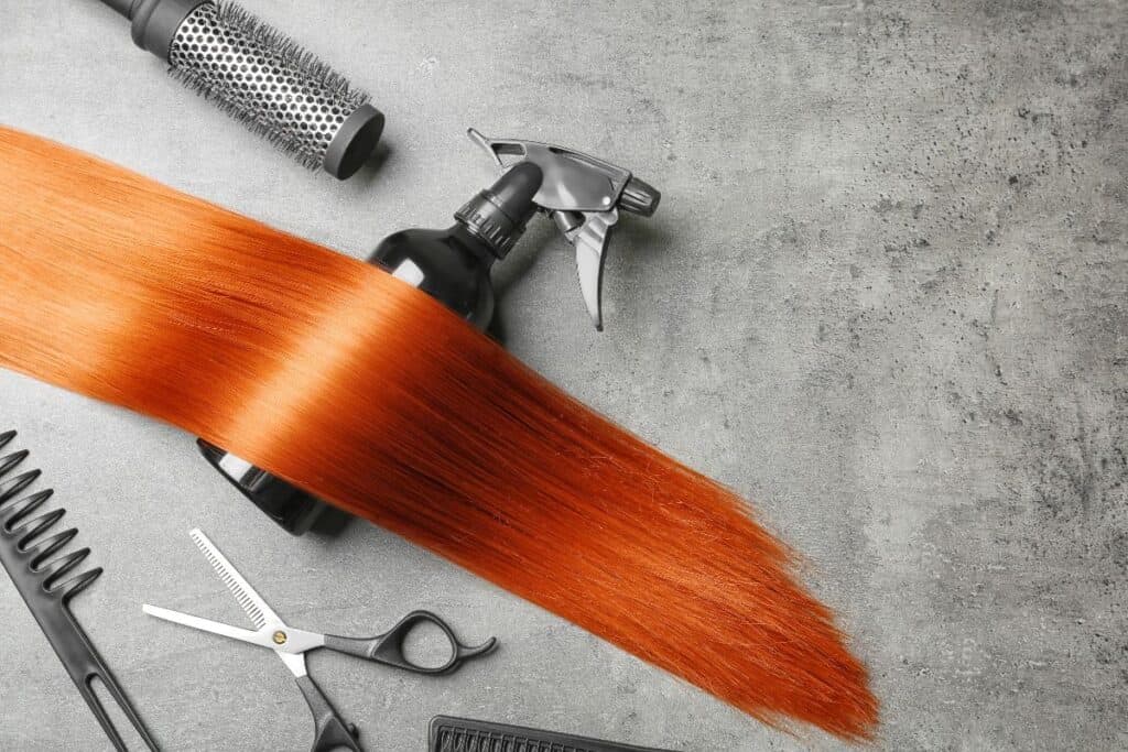 Copper hair weft with spray bottle, round brush, comb and scissors on a grey surface