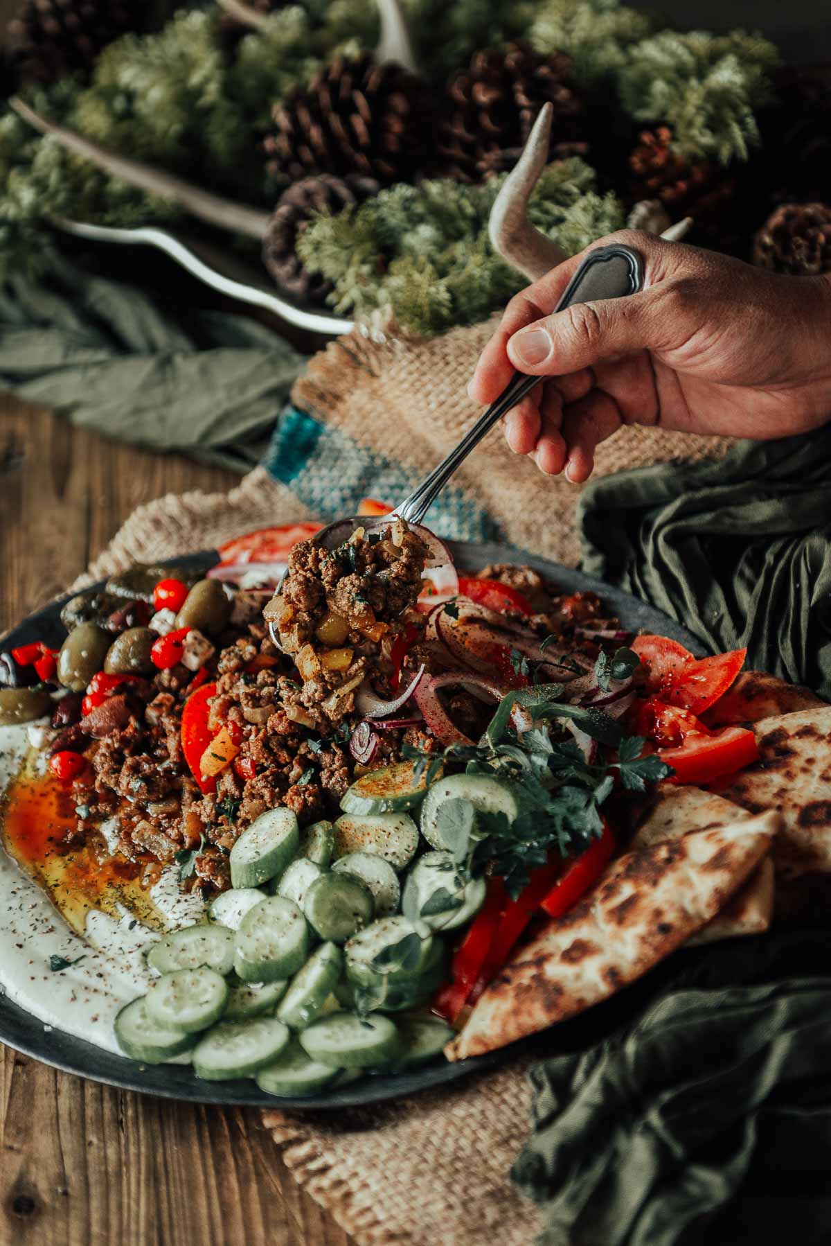 A hand holding a fork hovers over a plate with greek ground lamb, sliced tomatoes, cucumbers, olives, red onions, pita bread, and herbs on a rustic table setting.