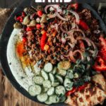 A plate of Greek ground lamb with sliced cucumbers, onions, tomatoes, olives, pita bread, and a whipped ricotta, displayed on a wooden table. Text reads "How to Make Greek Ground Lamb.