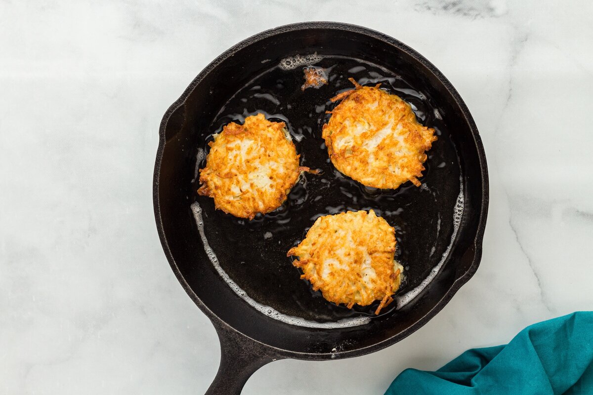 Latkes turning golden brown as they continue frying in a cast-iron skillet.