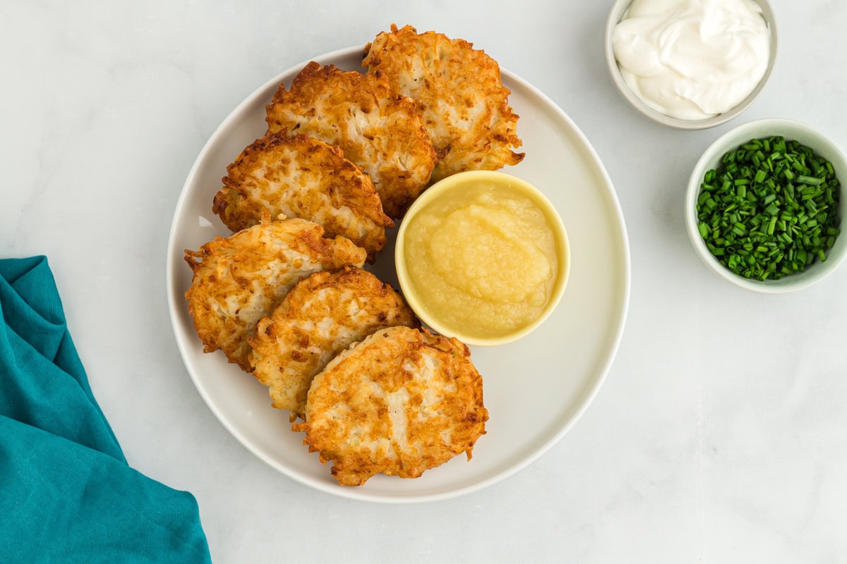 Plate of gluten-free potato latkes arranged around a bowl of applesauce, with sour cream and chives on the side for serving.
