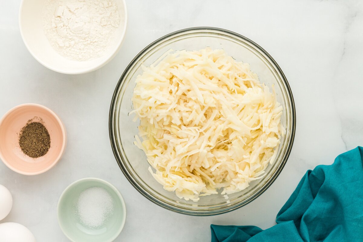 Large bowl of freshly grated russet potatoes with small bowls of gluten-free flour, salt, pepper, and eggs around it.