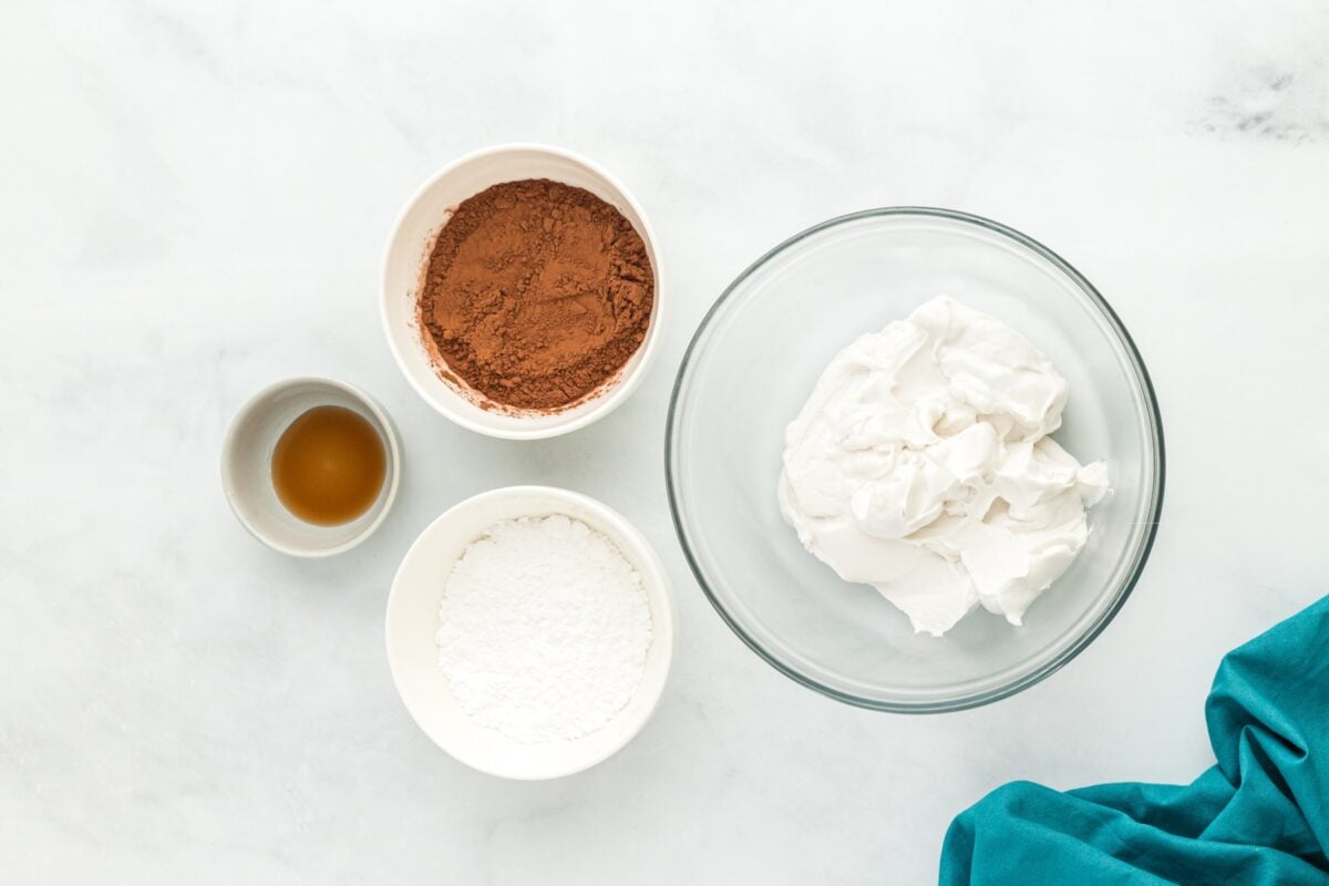 Bowls of coconut cream, cocoa powder, powdered sugar, and vanilla extract arranged on a white surface before making vegan chocolate mousse.