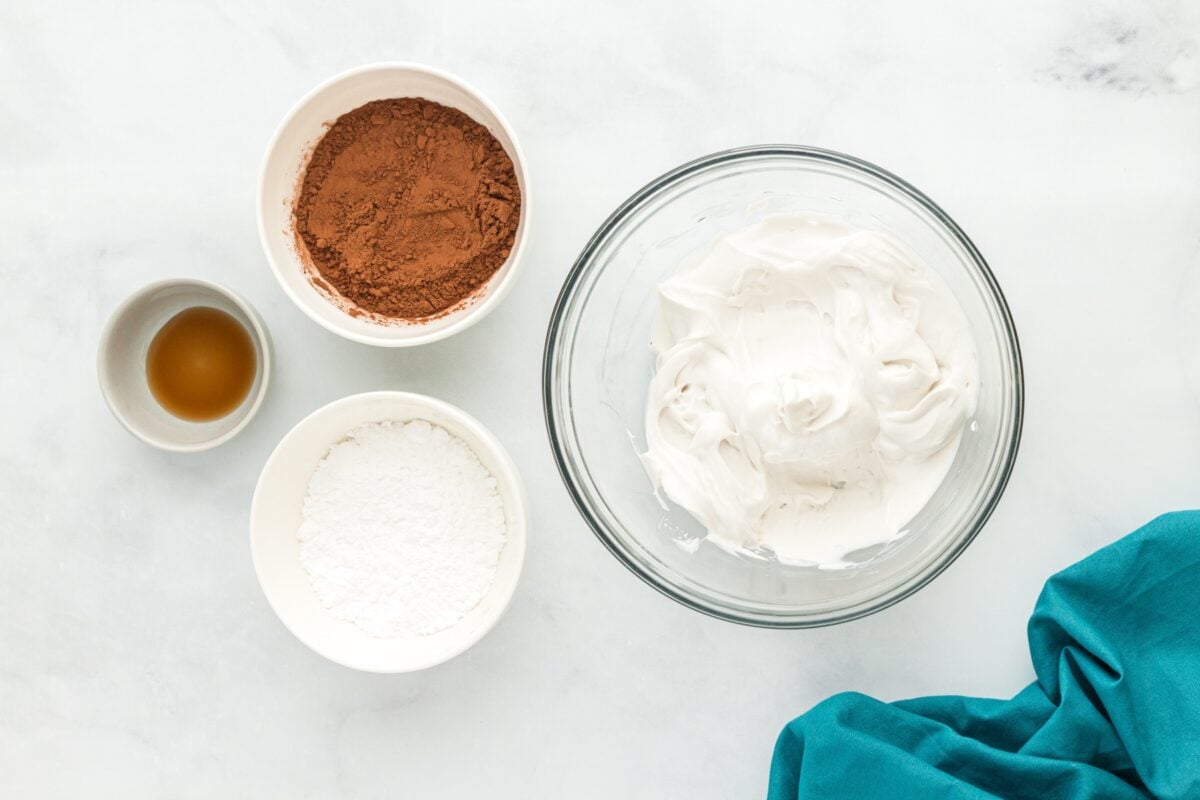 A mixing bowl filled with whipped coconut cream next to small bowls of cocoa powder, powdered sugar, and vanilla extract.