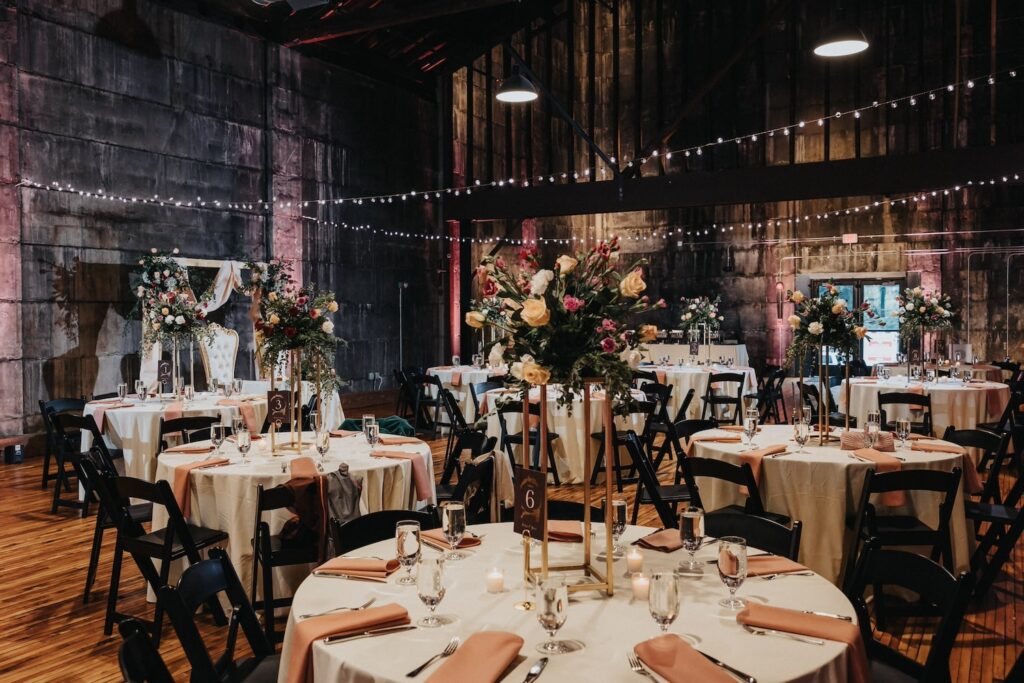 Round guest tables with tall floral centerpieces decorate a large, industrial event space
