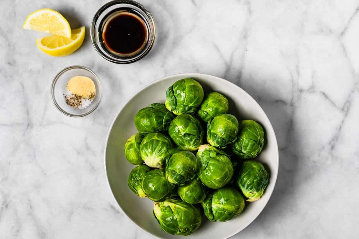 Raw Brussels sprouts in a clear bowl with two small bowls of balsamic vinegar, oil, and seasonings on a marble surface.