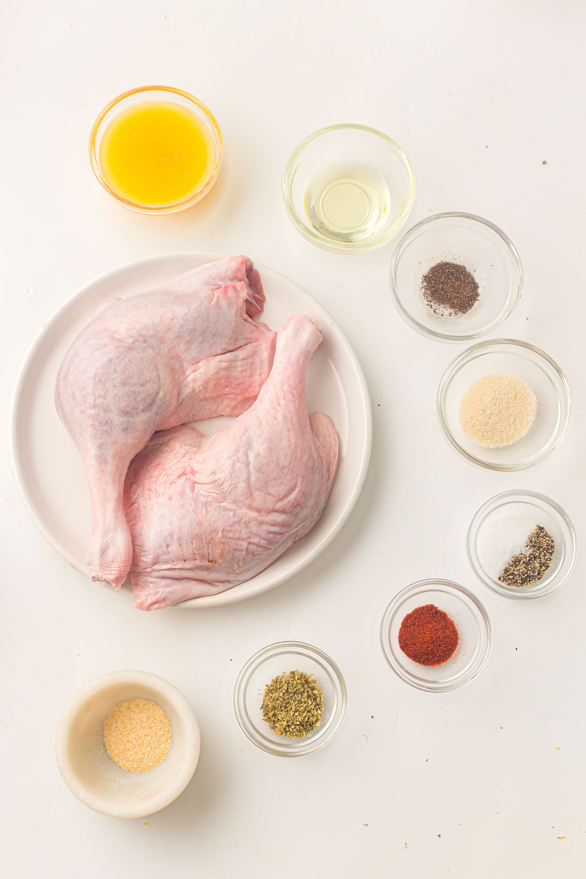 Two raw duck legs on a plate, surrounded by small bowls containing oil, orange juice, and various spices arranged on a white surface.