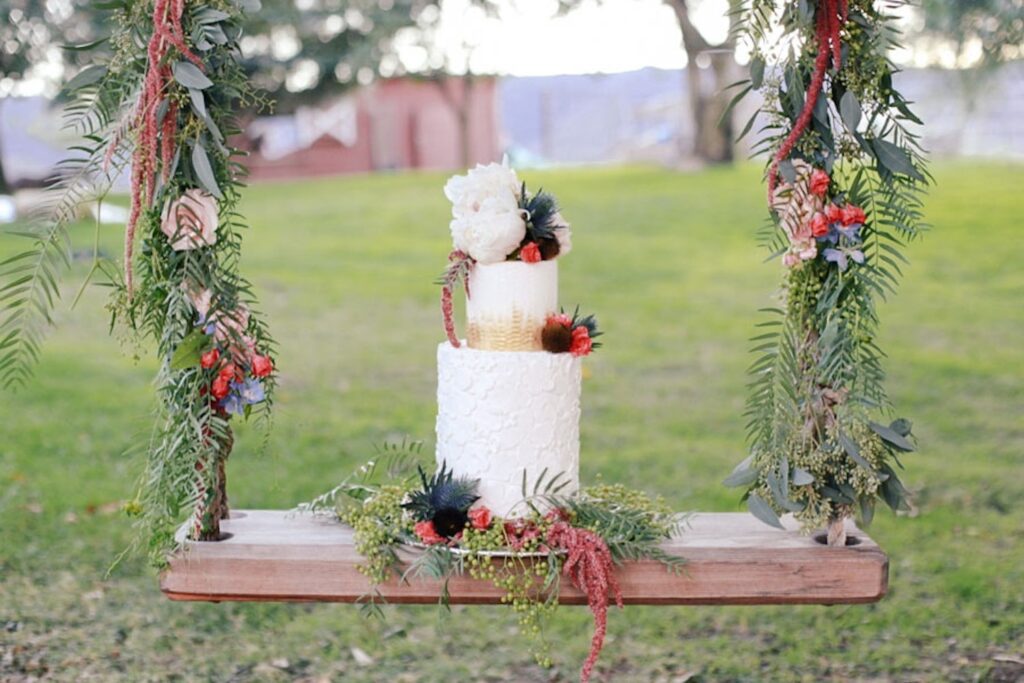 A white wedding cake with gold accents sits on a wooden swing draped in greenery and pink-hued florals