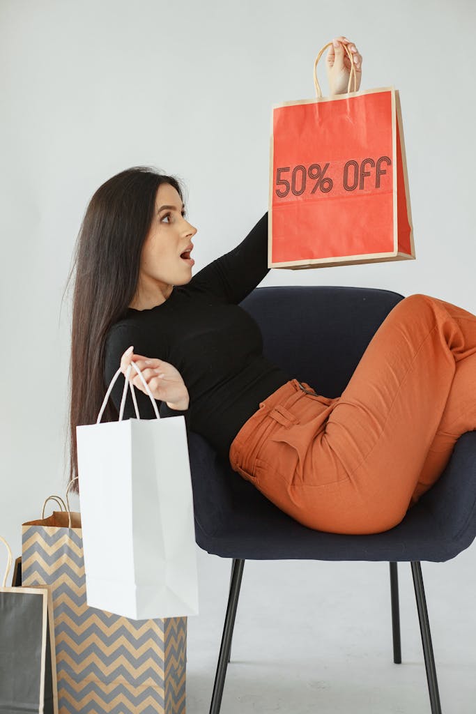 Woman sitting on chair holding shopping bags with discount, expressing excitement. Looking for sales is one way to travel solo on a budget